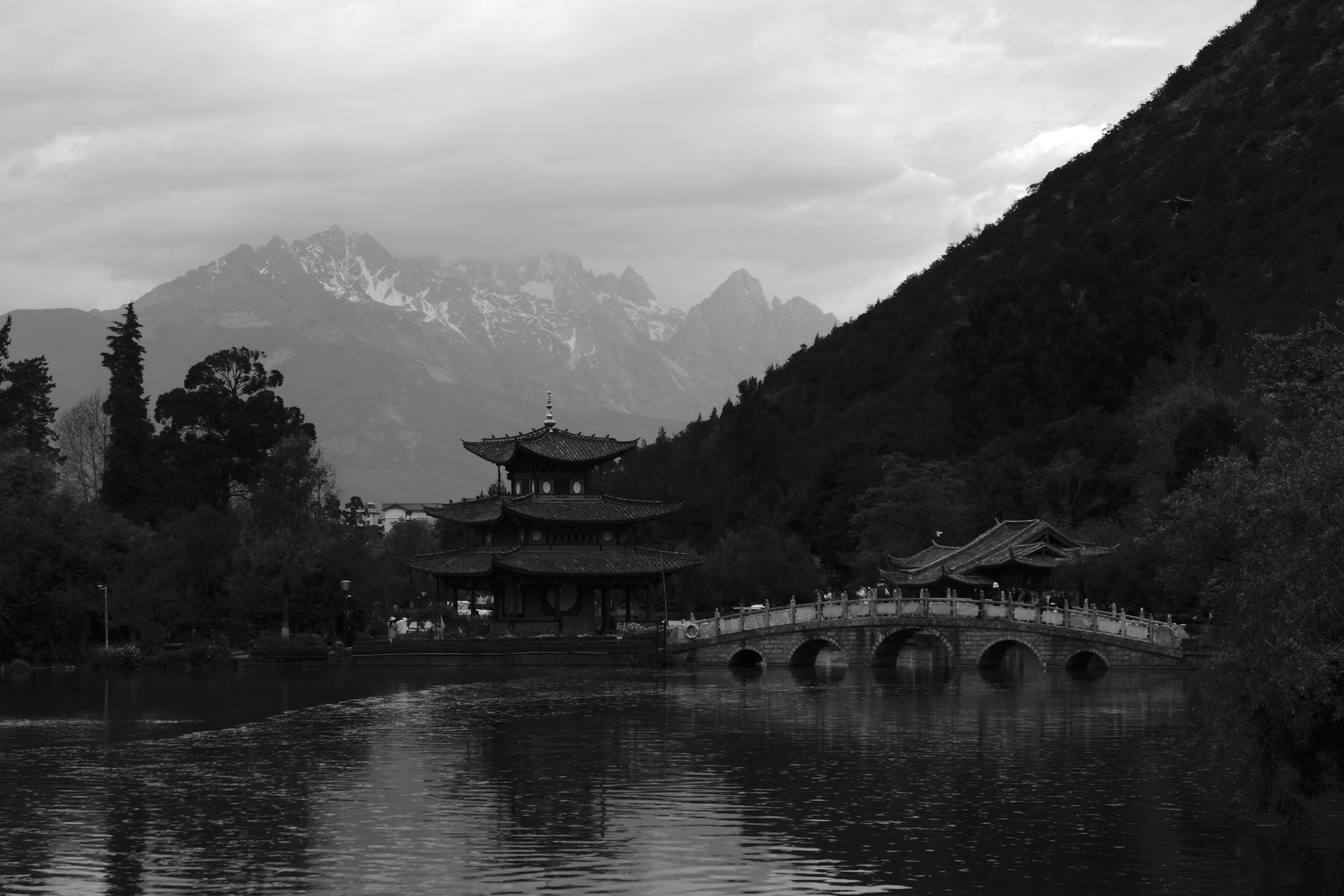 A Chinese temple by the side of a lake with an arched bridge leading up to it and both are reflected in the still water, in front of a snow covered mountain. A creative filter is applied to the image