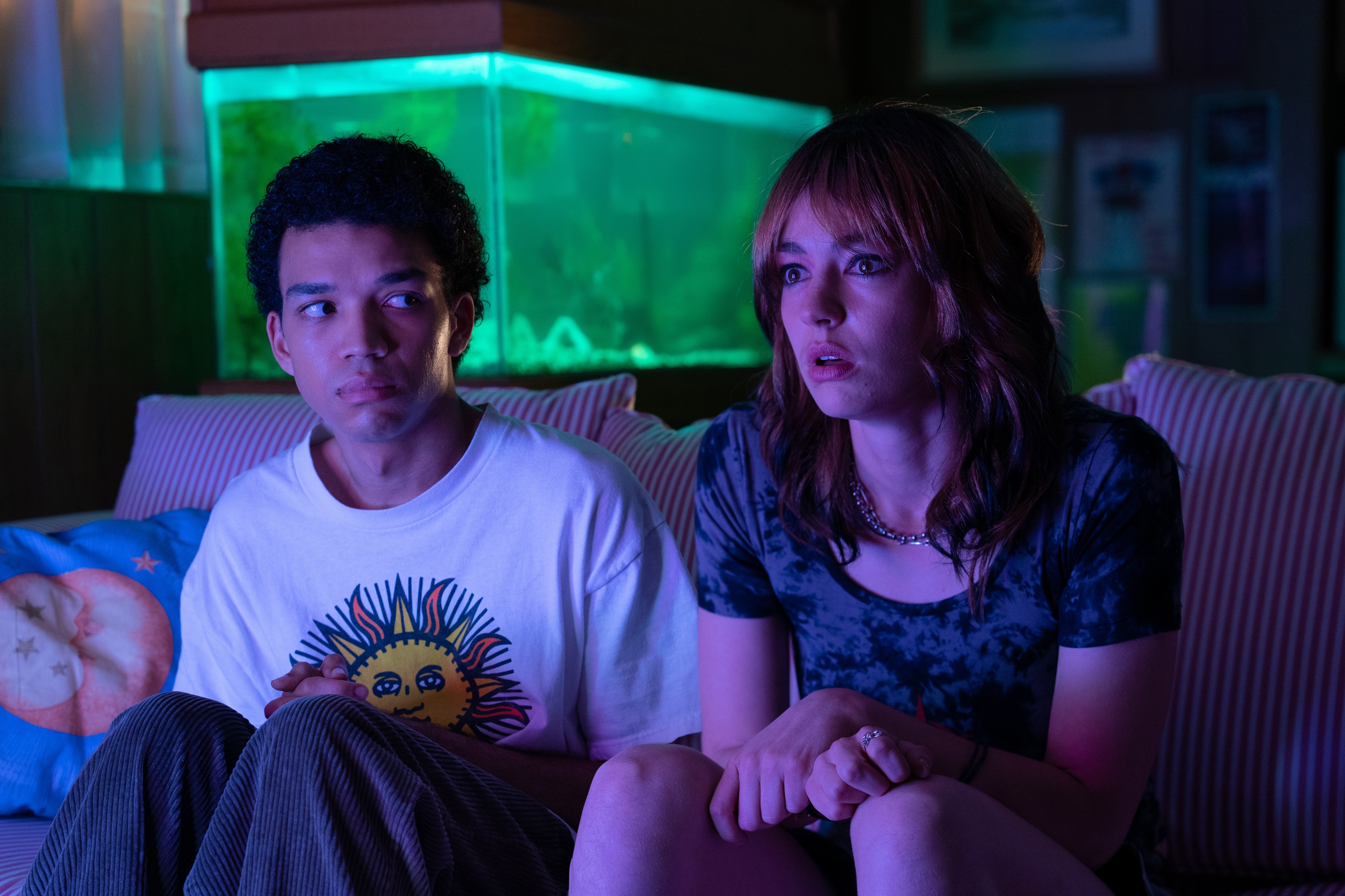 a man and a woman sit on a couch illuminated by the tv's glow, with a fish tank behind them