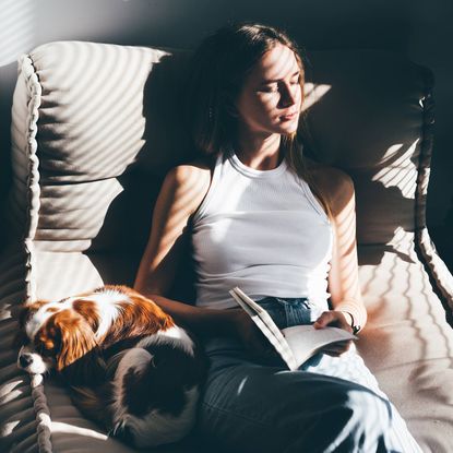 Woman reading a book and cuddling a dog on a sofa. 
