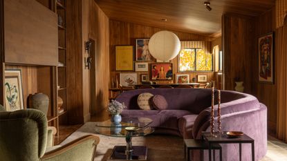 A mid-century living room with paneled walls, a sloped paneled ceiling, and curved purple couch, and collected artworks