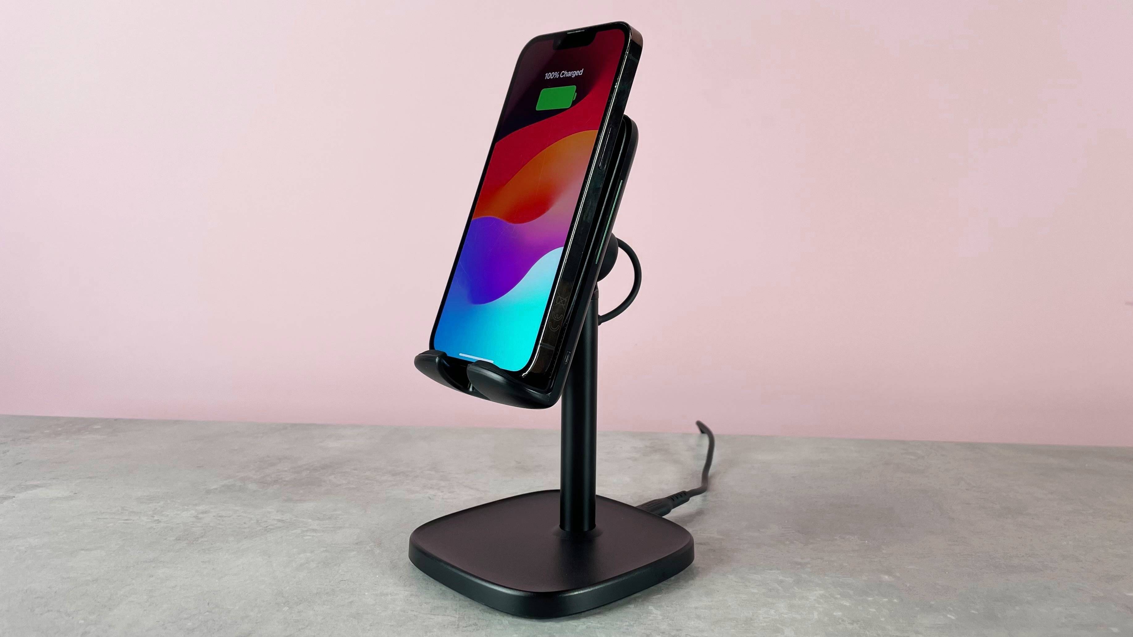 The INIU I212 Adjustable Wireless Charger is pictured sitting on a grey stone surface against a pink background. There is an iPhone on the charger; its screen shows that it is charged to 100%.