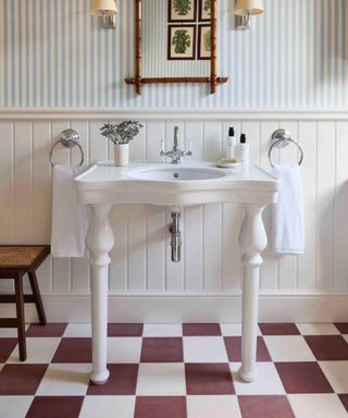 Bathroom with blue and white stripe wallpaper, white basin, shiplap, plum and white check floor tiles, rattan mirror, wall lights, towel rings
