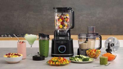 A Ninja blender filled with fruits; a Ninja food processor making a dip; surrounded by plates of cut vegetables and glasses of blended fruits, all on a white counter against a gray background. 