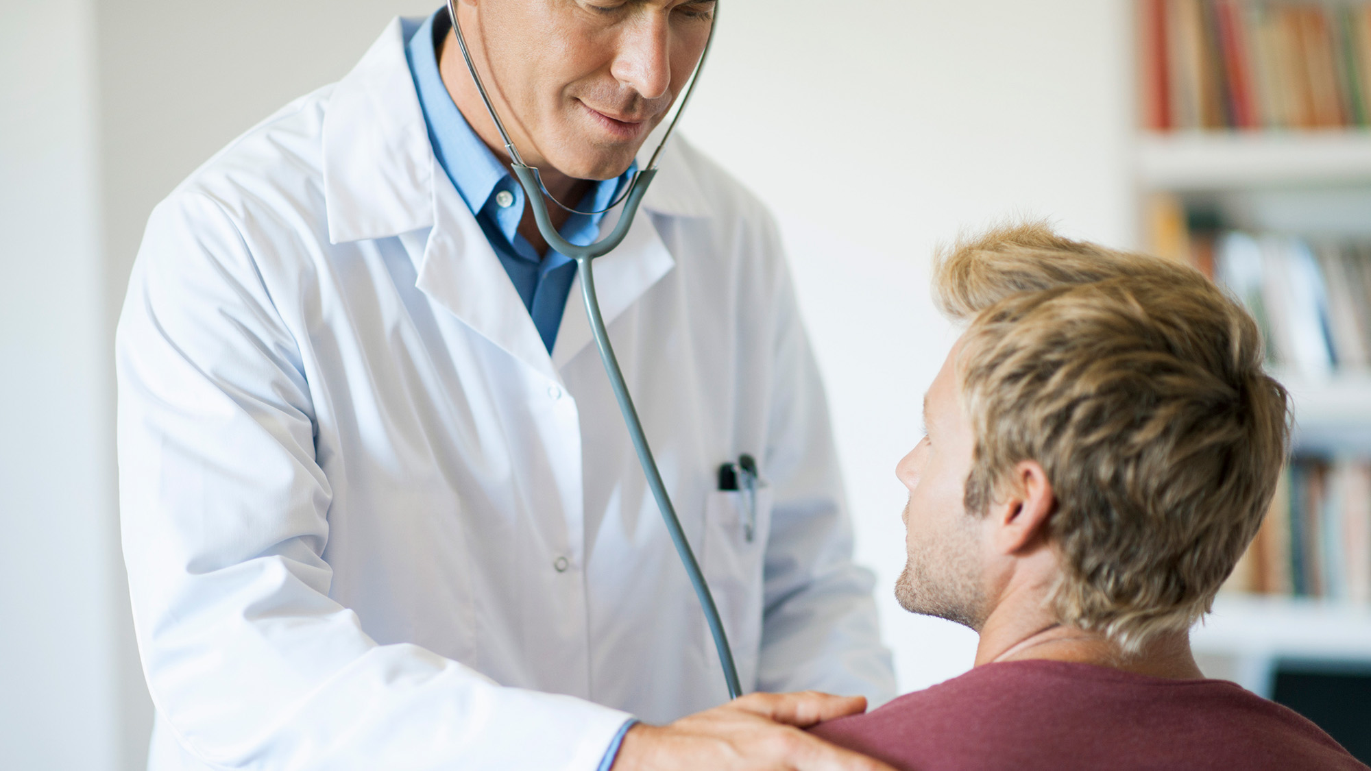 A man wearing a lab coat and stethoscope stands over a man in a maroon shirt