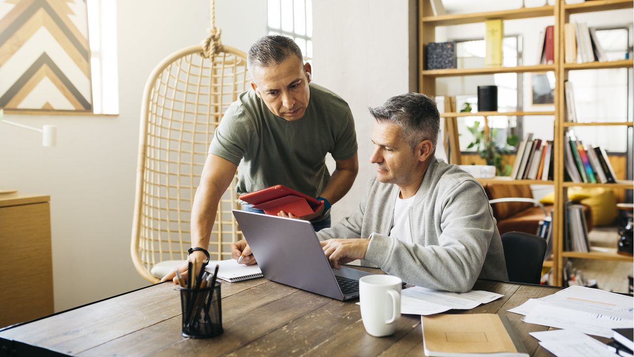 Two business owners look at a laptop on a desk, one sitting and one standing.