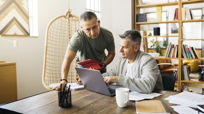 Two business owners look at a laptop on a desk, one sitting and one standing.