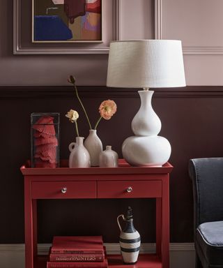 Light purple painted room with dark purple painted paneling, painted picture on wall, white lampshade, red side table with books and decorative ornaments, blue chair