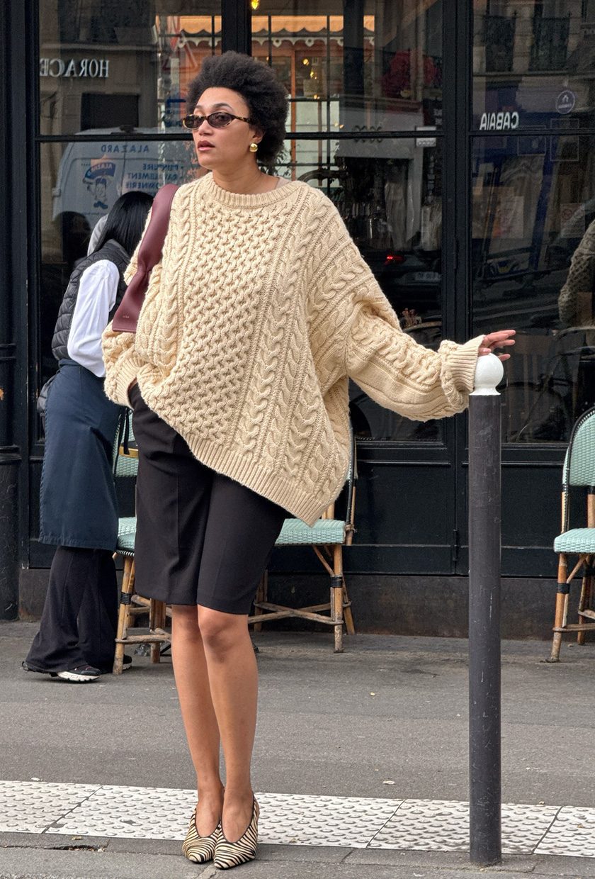 A woman wearing a camel cable-knit sweater with black pleated shorts, zebra-print pumps, and a red shoulder bag.