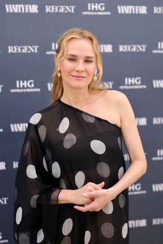 Diane Kruger smiling and wearing a sheer black and white polka dot one-shoulder gown, with her blonde hair in a half-up half-down hairstyle