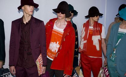 Six male models wearing looks from the Burberry Prorsum collection. One model is wearing a belted maroon piece with a maroon jacket over the top. Next to him is a model wearing a red jacket, dark coloured trousers and an orange and white t-shirt featuring a train design. Another model is wearing a dark blue coat. The fourth model is wearing a white and orange t-shirt, red sleeveless V-neck top, red trousers and a red and white scarf. The fifth model is wearing a light brown coat. And the sixth model is wearing a blue and white t-shirt, blue trousers and a blue cardigan. All models are wearing hats, three are wearing sunglasses and some are holding briefcases and notebooks