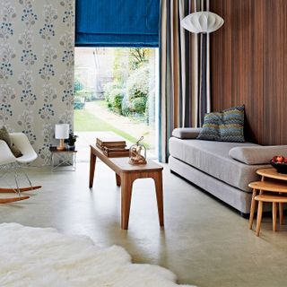grey John Lewis Sonoma sofa bed to the right against a wooden slat wall, patterned floral wallpaper to the left next to a wide window with blue blind, and slate tile floors, with a wooden coffee table and large white rug