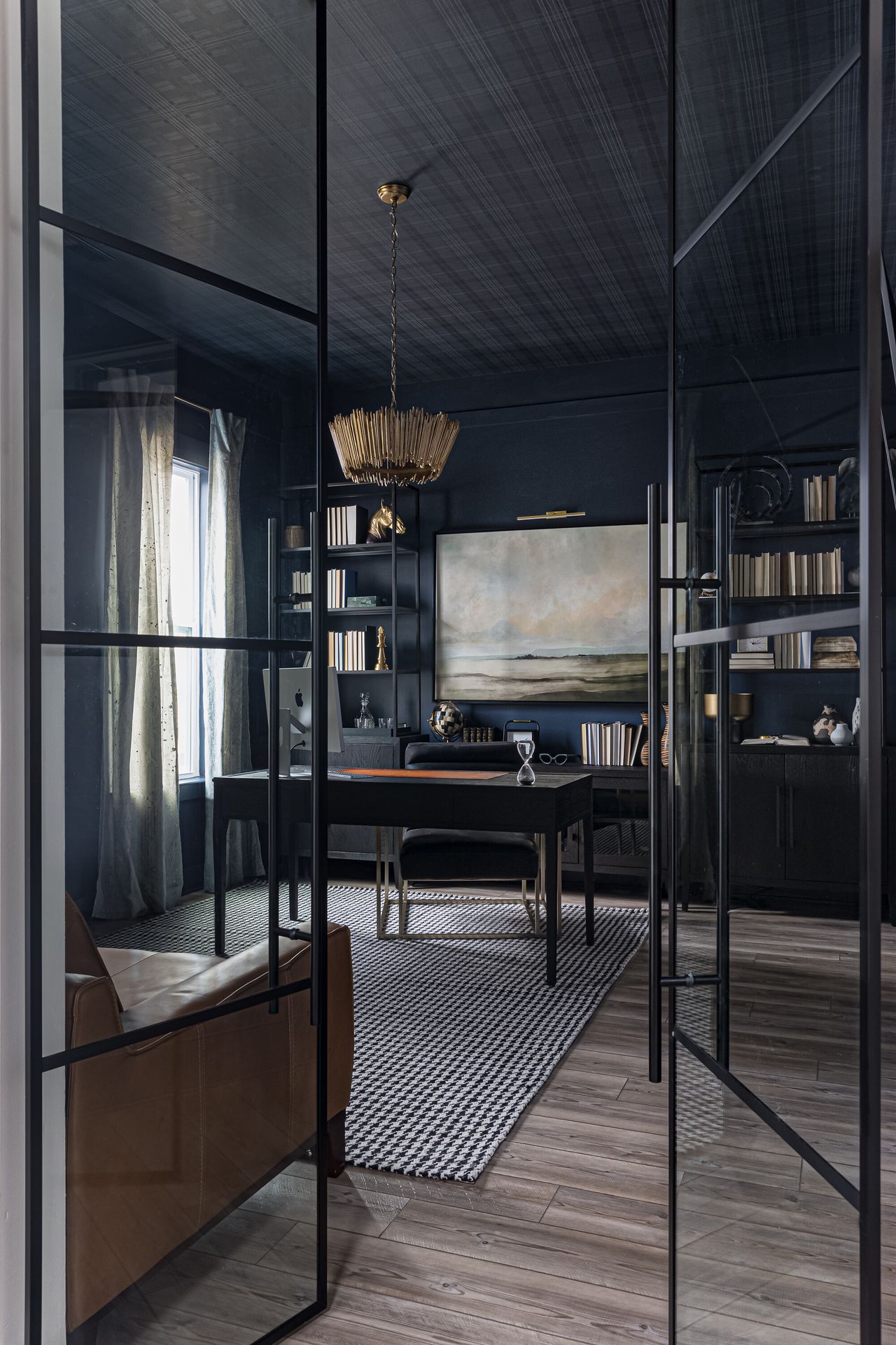 Image of a traditional style home office that is painted in a navy blue paint color. There is a slim black desk and black desk chair and two slim black bookshelves on either side of the back wall. There is a light, landscape painting in between the shelving units.