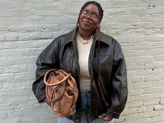 Chichi wears brown bomber, white tank, and denim jeans while holding a light brown leather tote bag