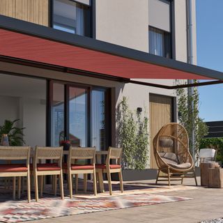 Orange awning with black frame on the side of a grey painted contemporary house, extended out over a patio with wooden dining table