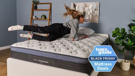 A woman jumping on the Helix Midnight Luxe mattress on white bedframe in sleep studio with blue wall, bedside shelving on the left and plants on the right