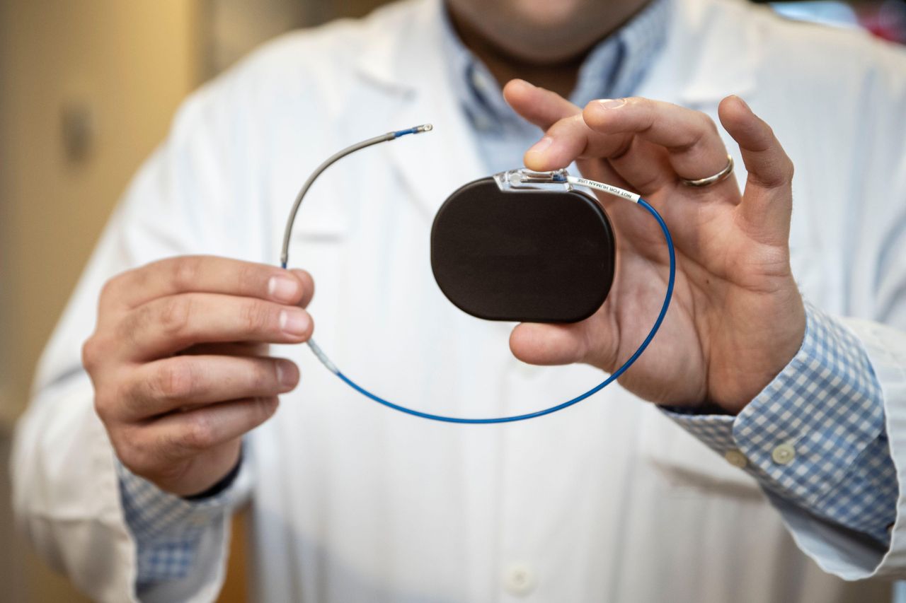 A medium shot of the hands and torso of a person in a white lab coat. They are holding a palm-sized black device with a wire attached to the top.
