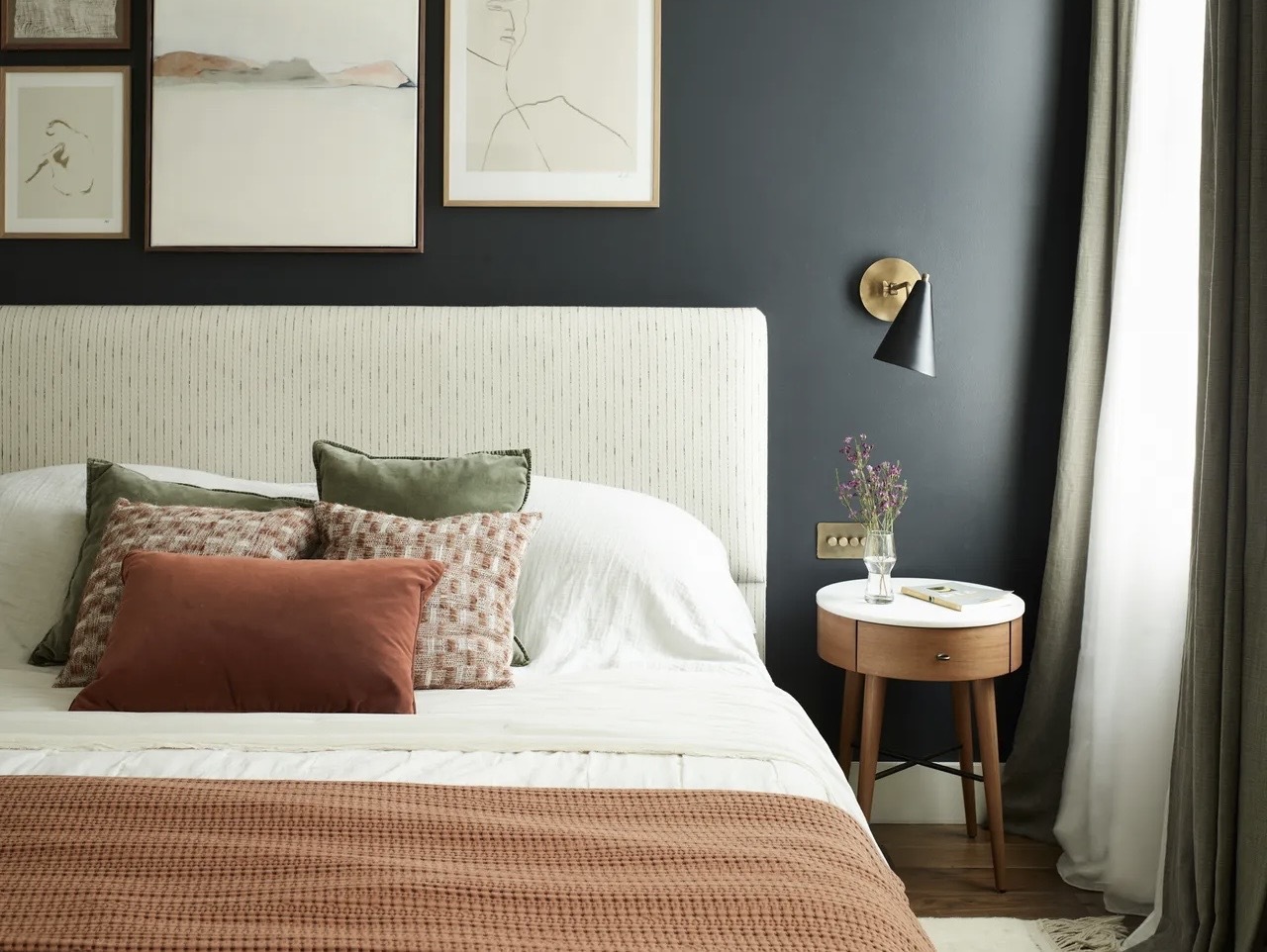 A bedroom layered with cushions in a nature-inspired palette infront of an inky blue wall with artwork and a round wooden bedside table