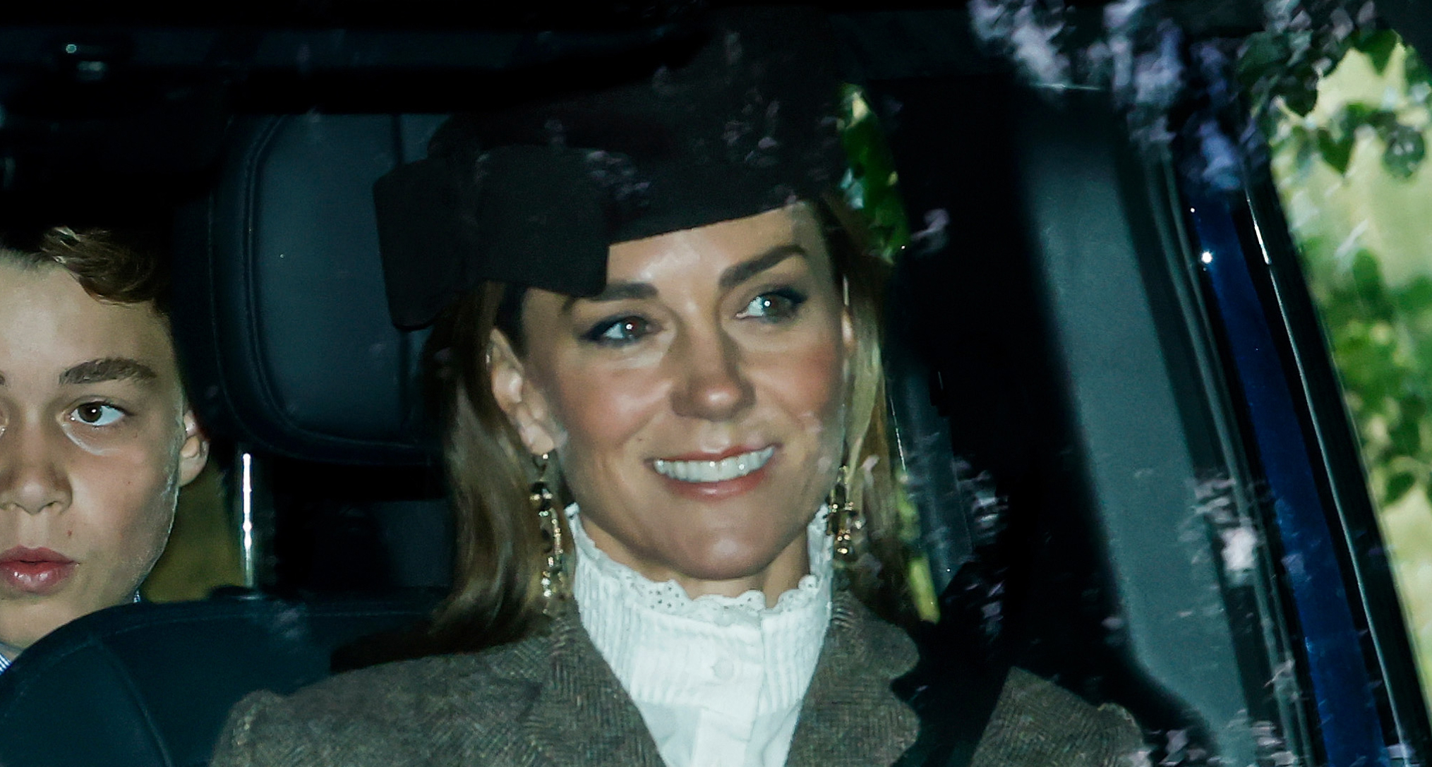 Princess Kate sitting in a car wearing a white blouse and brown hat