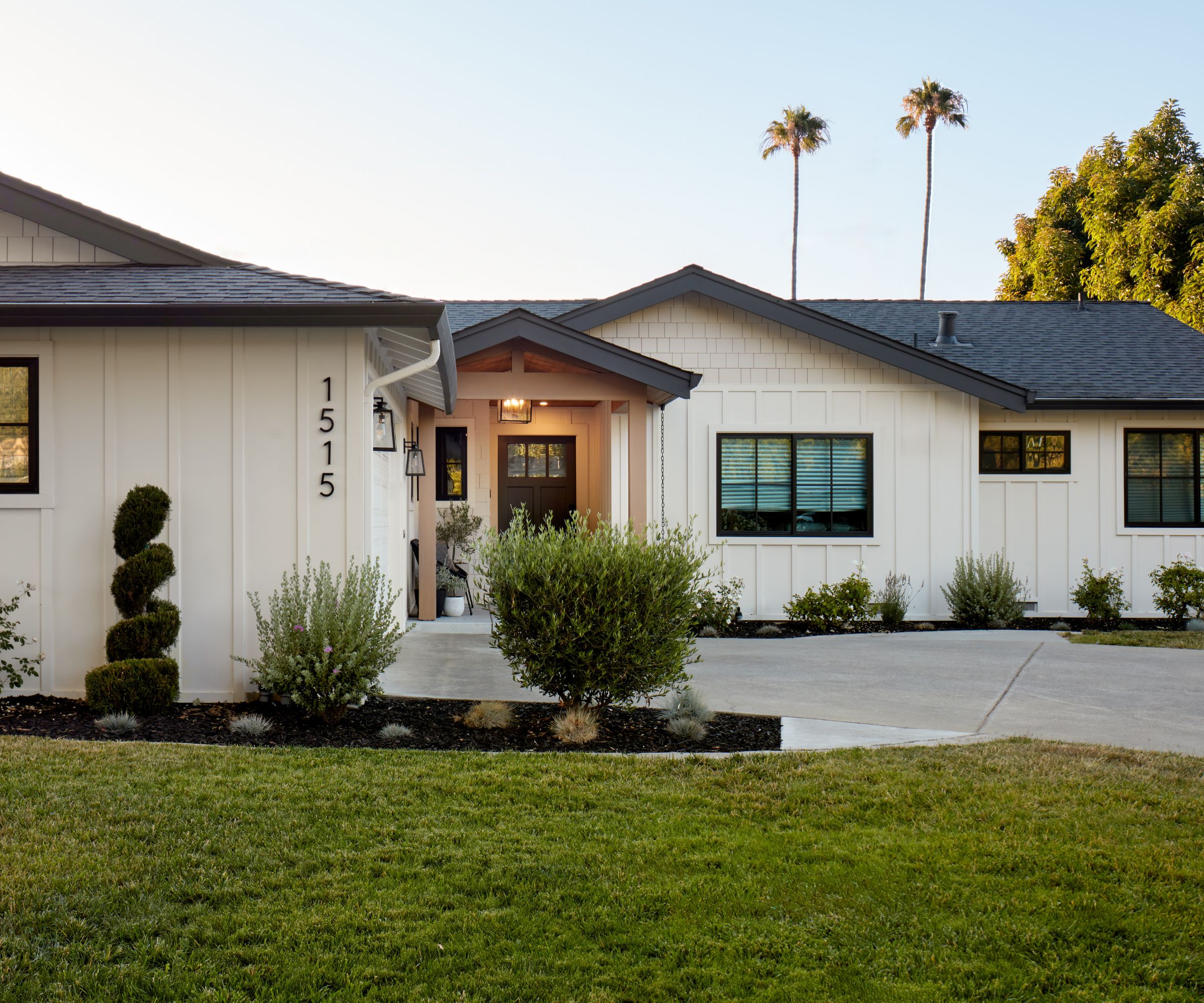 A bungalow home exterior with warm white siding, a dark brown door, and a light brown porch. Dark window trim and a gray roof with a grass lawn and paved driveway.
