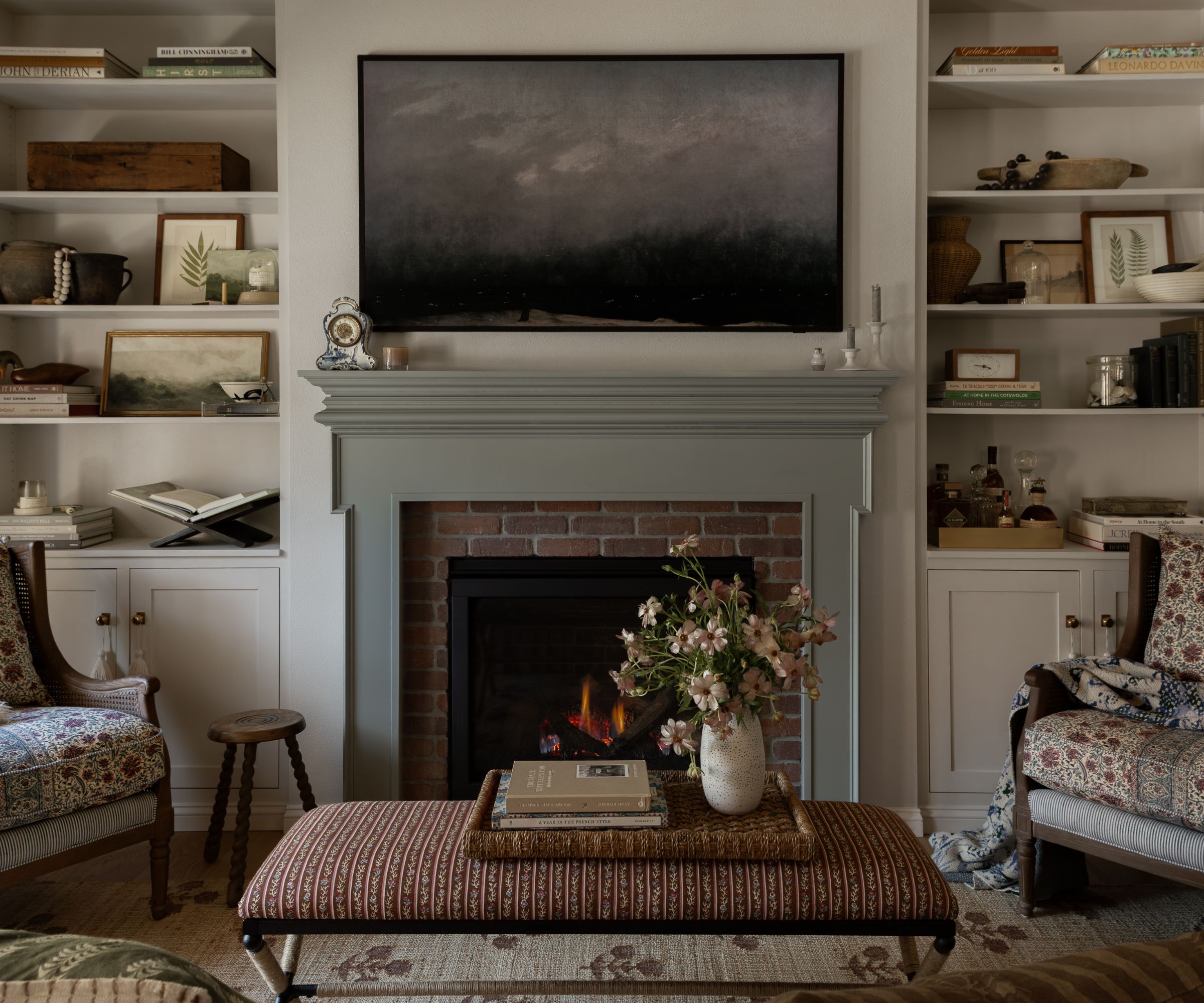 Cozy living room with soft white walls and built-ins with collected decor on show. A stone-colored fireplace with a landscape artwork above it. Two armchairs with a ditsy floral pattern and an ottoman