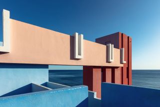 View of the pink, blue and white geometric wall arrangement at La Muralla Roja by Ricardo Bofill in Calp, Spain. The sea can partially be seen and the sky is clear and blue