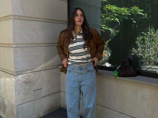 Borislava wears brown suede jacket, rugby shirt, wide jeans, and dark close-toed shoes while posing for the camera outside.