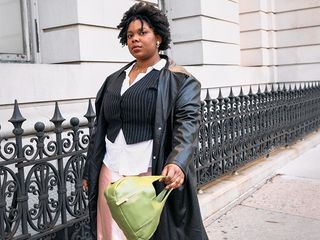 Chichi wears a layered loo consisting of a satin dress, button down shirt, vest, and faux leather trench, and a green purse