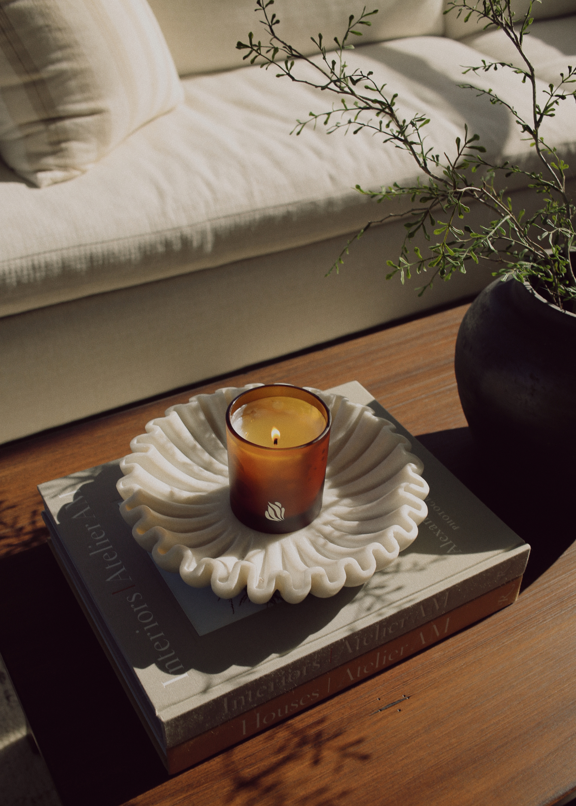 A scented candle from Nose Dive on a textured stone bowl on a stack of books by a vase of stems on a coffee table
