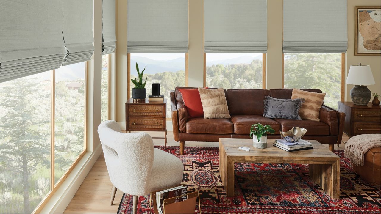 Small living room with windows revealing trees and green space outside, partially covered by gray blinds. Pictured in the living room are a brown sofa, a white armchair, wooden drawers and coffee table, and a red patterned rug.