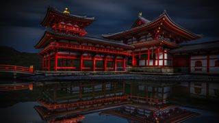 A dramatic, low-light photograph of the red and white wooden structures of the Byōdō-in Temple in Uji, Japan, reflected clearly in the dark water of a pond, with a single white koi fish near the bottom left corner.
