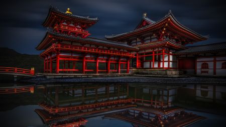 A dramatic, low-light photograph of the red and white wooden structures of the Byōdō-in Temple in Uji, Japan, reflected clearly in the dark water of a pond, with a single white koi fish near the bottom left corner.