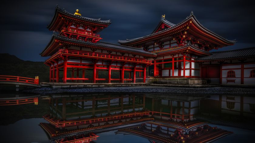 A dramatic, low-light photograph of the red and white wooden structures of the Byōdō-in Temple in Uji, Japan, reflected clearly in the dark water of a pond, with a single white koi fish near the bottom left corner.