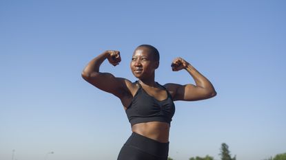 A woman flexes her muscles and smiles against a blue-sky background.