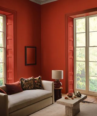 modern living room with brick red walls and shutters, taupe couch and cream rug, stone coffee table, artwork, lamp, patterned couch pillows