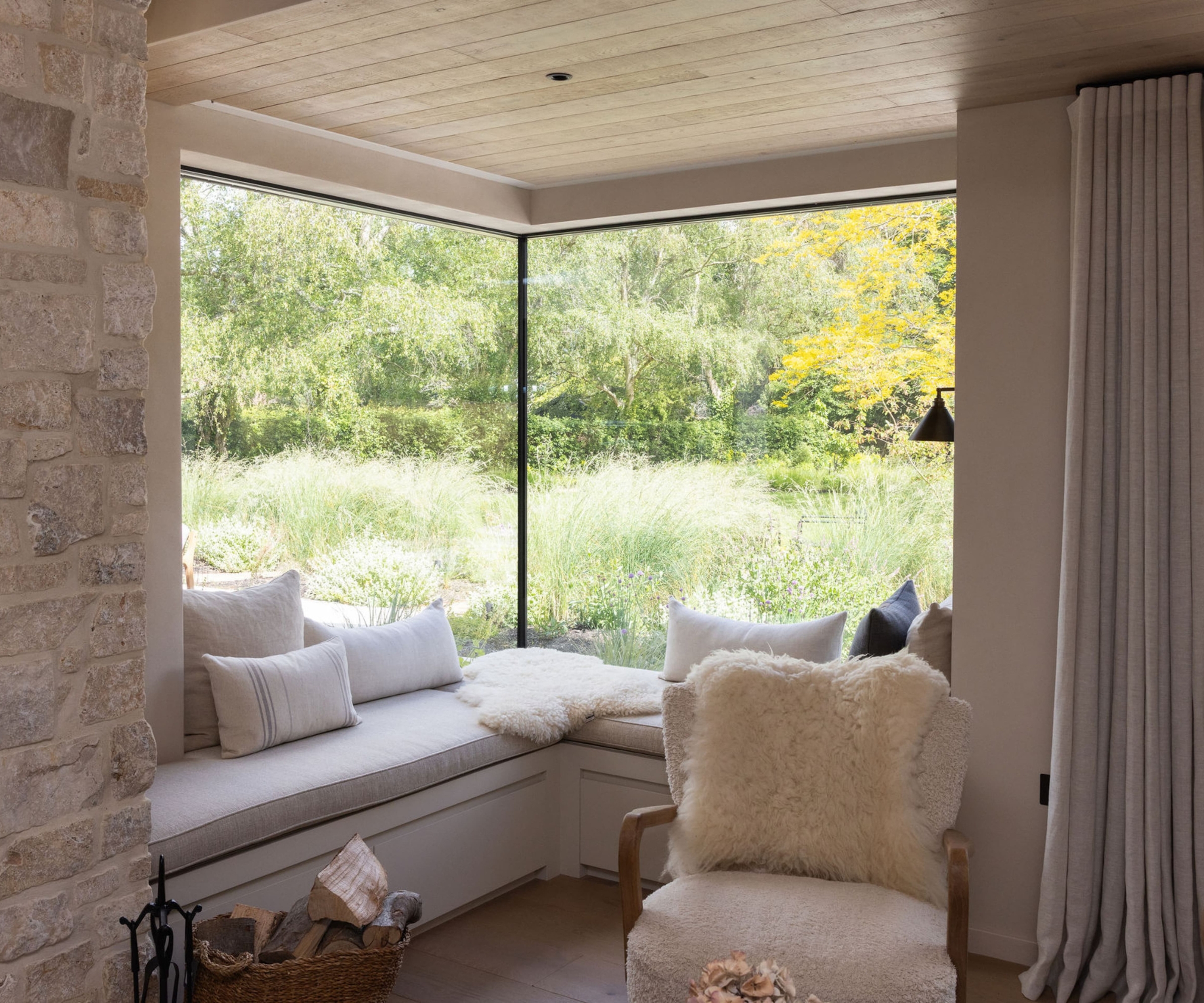 Cosy corner window seat with cushions overlooking a wildflower meadow, beside a sheepskin-draped armchair.