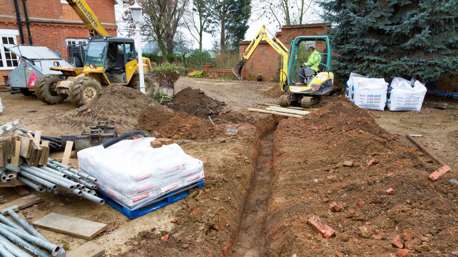 A digger digging a trench for drainage with a red brick house in the background and pipes in the foreground
