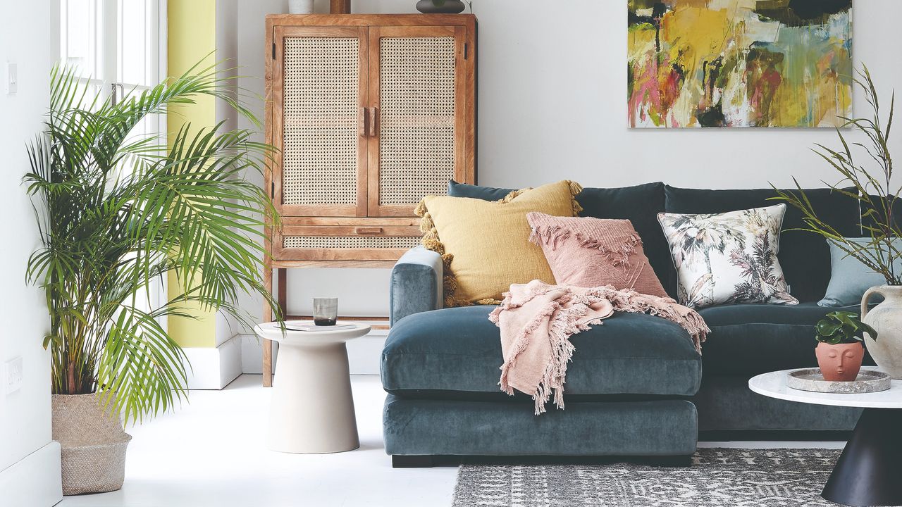 A living room with a dark blue velvet corner sofa, an expressive abstract artwork hanging on the back wall and a rattan-insert cabinet