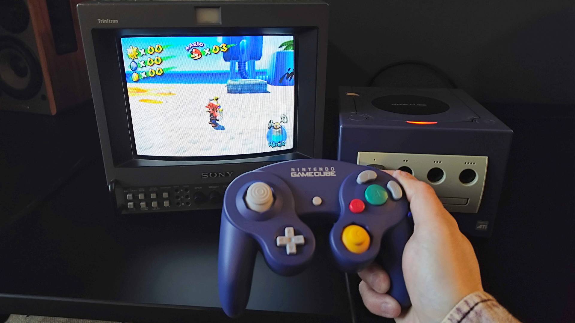 Hand holding Switch 2 GameCube controller in front of original console and Sony PVM CRT with Super Mario Sunshine gameplay on screen.
