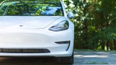  An all electric Tesla Model 3 in white on cement road with trees in background on sunny day.