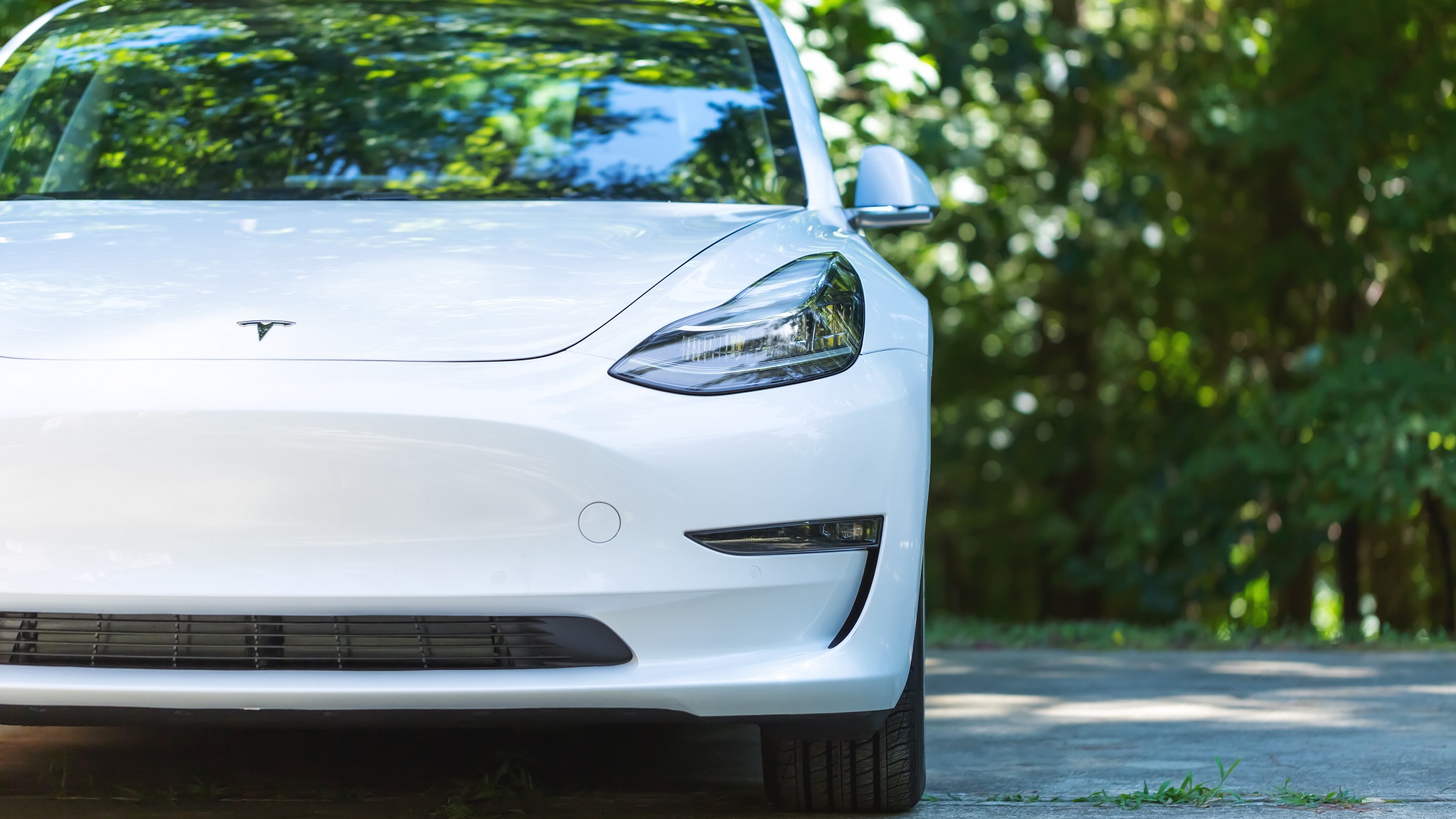 A white all-electric Tesla Model 3 on a cement road with trees in the background on a sunny day.