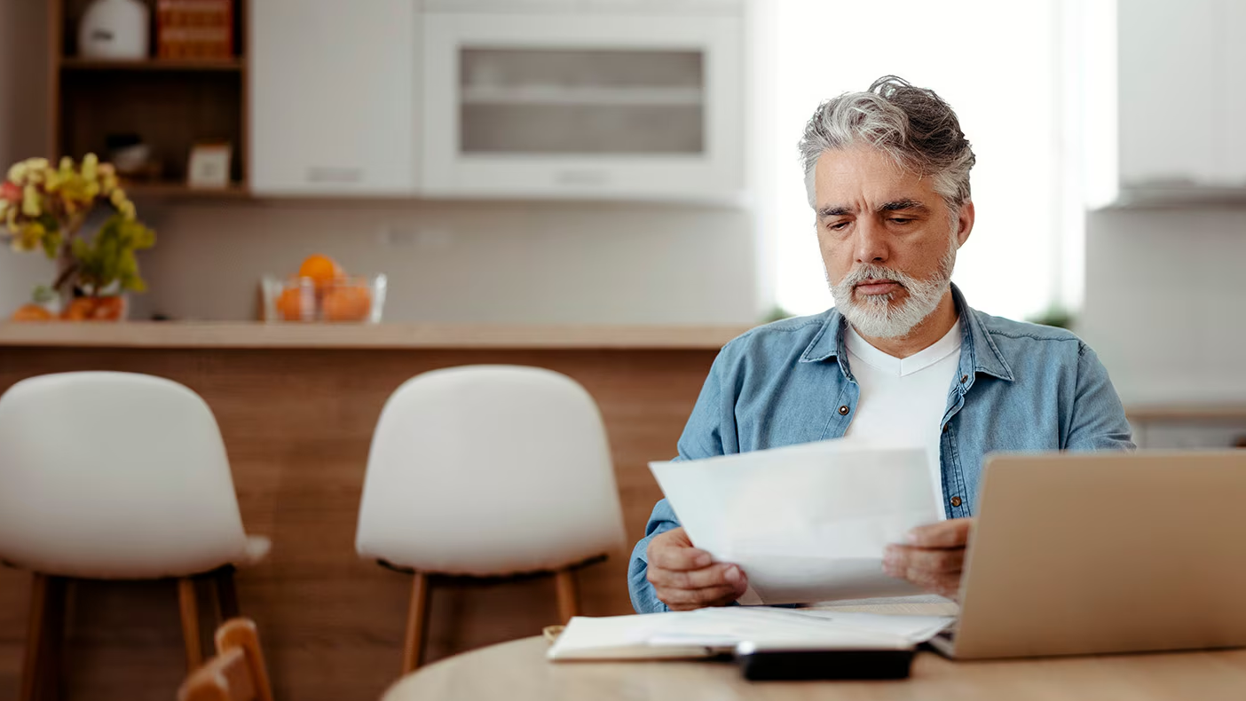 A man examining a piece of paper in front of a laptop.