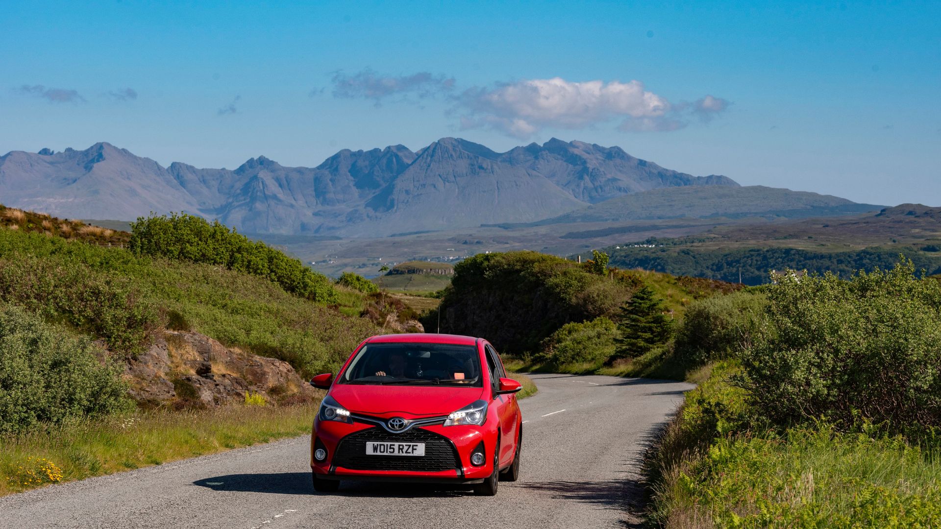 an image of a red toyata yarris driving along Isle of Skye, Scotland, UK. In the background is a scenic countryside view across to the Cuillin Hills with a blue sky