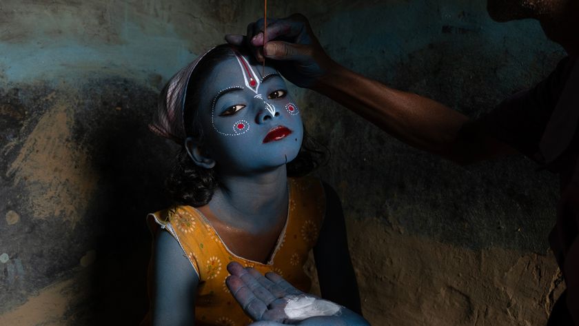 A child with blue skin sits calmly as an adult applies paint to their face in a dimly lit room