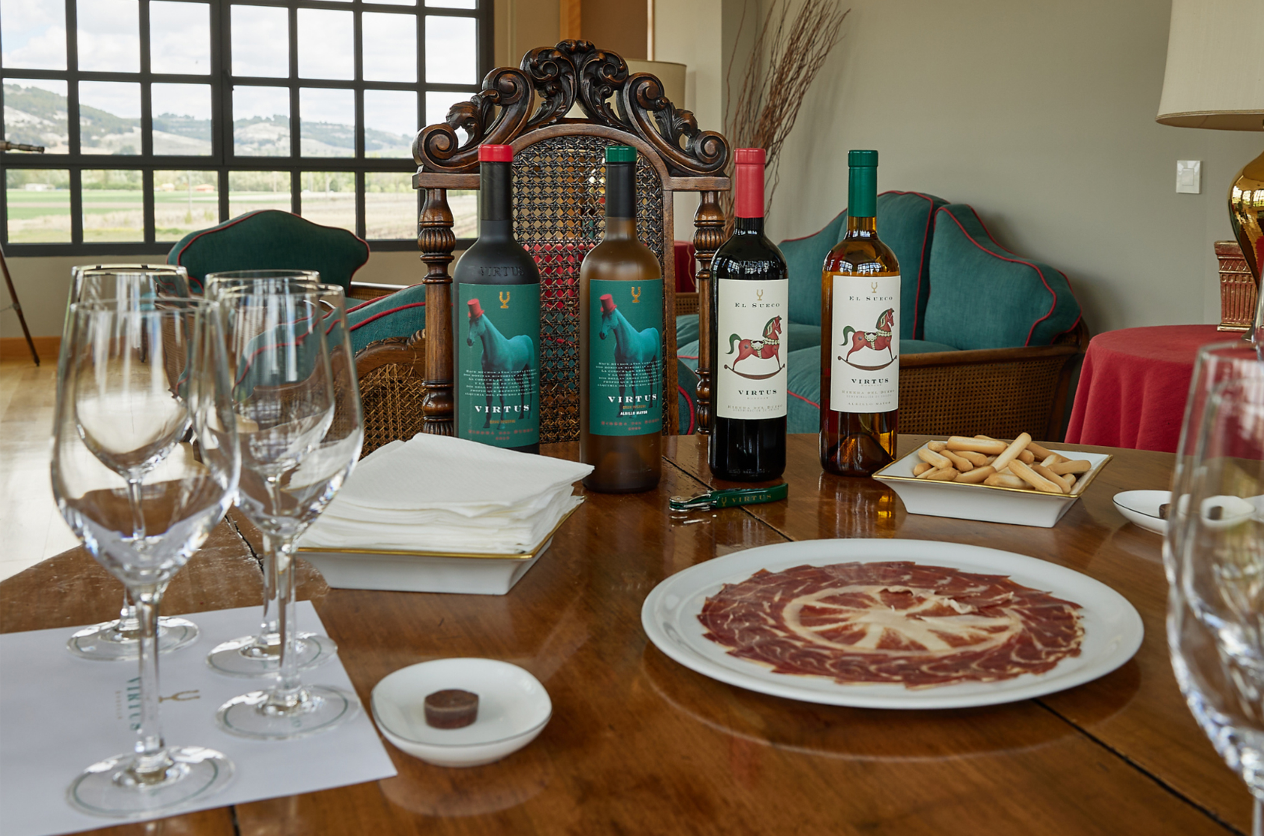 A wooden table with four different bottles, two red and two white, of Bodegas Virtus wines, with wine glasses and plates of snacks