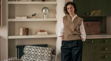 A young woman in a sand vest, a white shirt, and blue trousers poses in front of a built-in shelving unit with collectibles, next to a patterned sofa and a green chest of drawers, respectively.