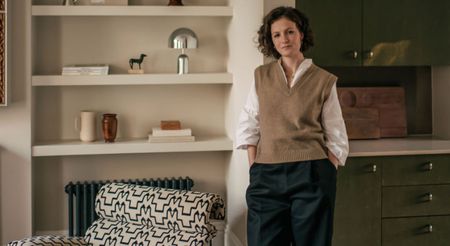 A young woman in a sand vest, a white shirt, and blue trousers poses in front of a built-in shelving unit with collectibles, next to a patterned sofa and a green chest of drawers, respectively.