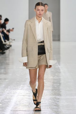 How to wear a suit is shown in a photo of a model on the runway of Max Mara's spring 2025 runway show wearing a light tan linen blazer over a white button-down shirt styled with a matching tan linen shorts, a dark brown belt bag, and dark brown flat sandals