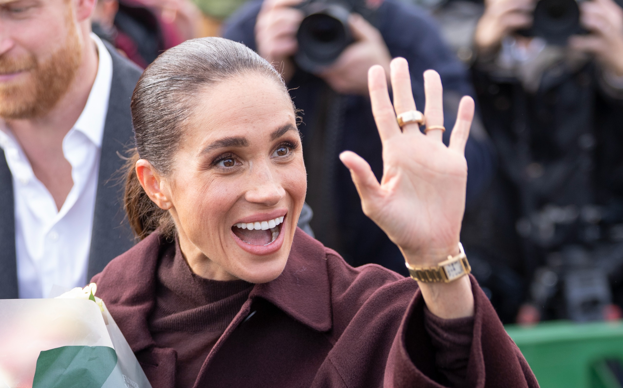 Meghan Markle waving and smiling in a maroon ncoat
