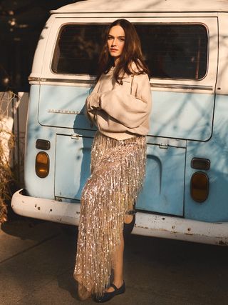 Emanuela Postacchini stands in front of a vintage van waring a cream sweatshirt and sequin skirt with black Mary Janes.