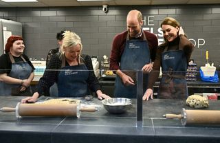 Kate Middleton and Prince William wearing blue aprons at a bakery laughing as a woman shows them how to roll out dough